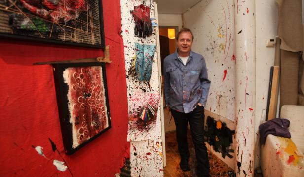 Ein Mann steht in einem Raum, der mit Farbspritzern bedeckt ist, neben einigen Kunstwerken.
