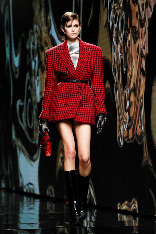 Ein Model in einem rot-schwarzen Hahnentritt-Blazer, schwarzen Stiefeln und Handschuhen auf einem Laufsteg.