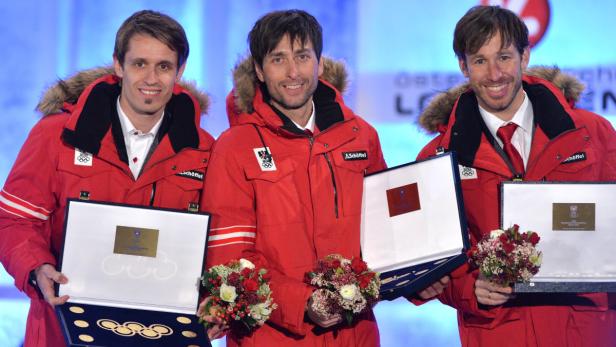 Drei österreichische Sportler in roten Jacken halten Auszeichnungen und Blumensträuße.