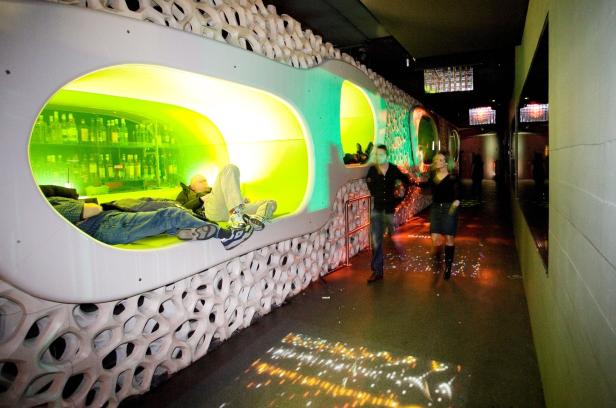 In einer futuristischen Bar entspannen sich zwei Männer in beleuchteten Nischen, während andere vorbeigehen.
