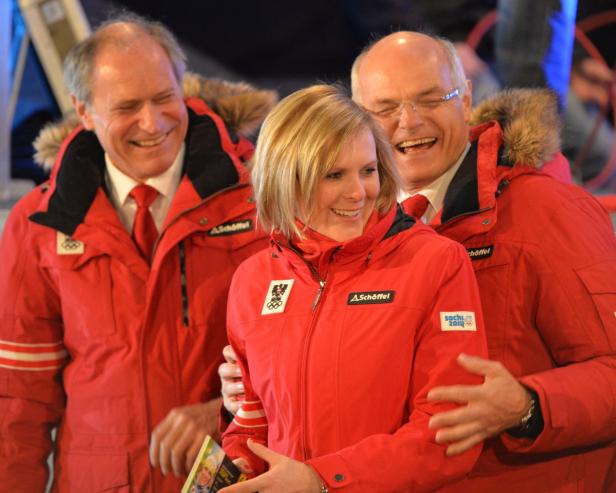 Eine blonde Frau in roter Jacke wird von zwei Männern in roten Jacken umarmt.