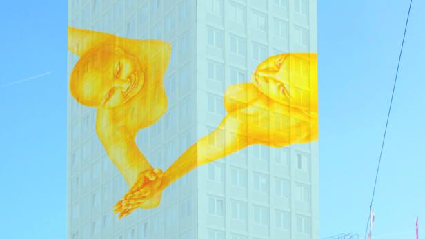 Detail aus dem Gemälde 'Verbundenheit' Ein Hochhaus mit einem goldenen Wandbild von zwei sich berührenden Figuren vor blauem Himmel.