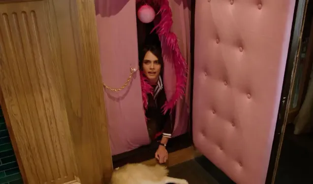 Cara Delevingne schaut aus einer rosa dekorierten Kabine, ein Hund liegt davor.