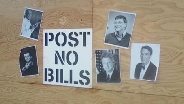 Ein Schild mit der Aufschrift „Post No Bills“ ist mit Porträts von Bill Gates, Bill Clinton und anderen beklebt.