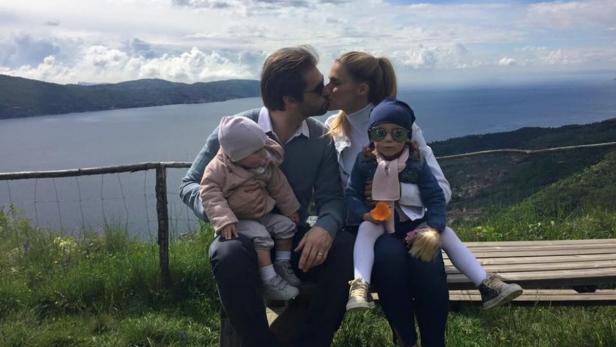 Tomaso und Michelle mit Sole und Celeste Eine Familie mit zwei kleinen Kindern sitzt auf einer Bank mit Blick auf einen See.