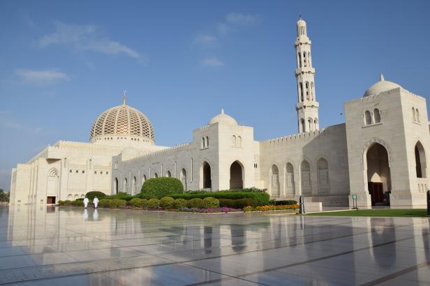 Weiße Moschee mit Kuppel und Minarett vor blauem Himmel, davor spiegelnder Platz und Blumenbeete.