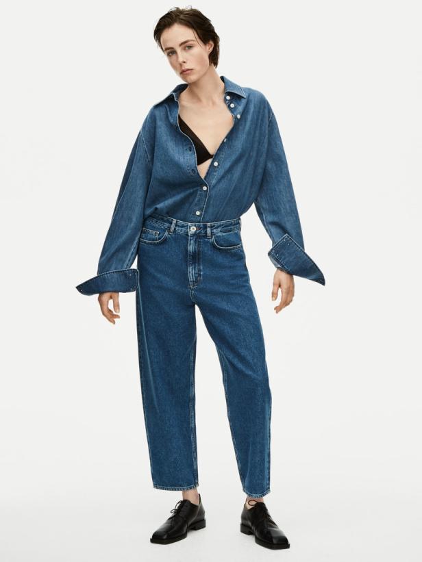 Eine Frau in einem Jeanshemd und einer Jeanshose posiert vor einem weißen Hintergrund.