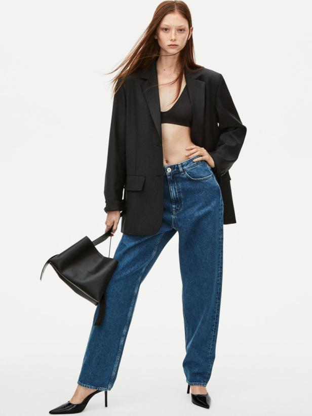 Eine Frau mit Blazer, Jeans und Handtasche posiert vor einem weißen Hintergrund.