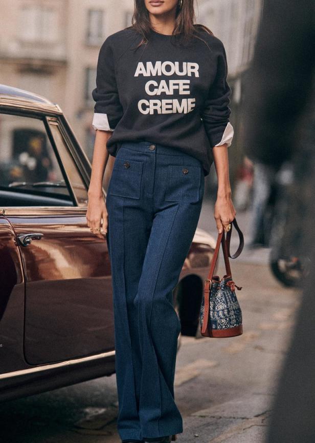 Eine Frau in einem Sweatshirt mit der Aufschrift „Amour Cafe Creme“ und Schlaghosen steht neben einem Auto.