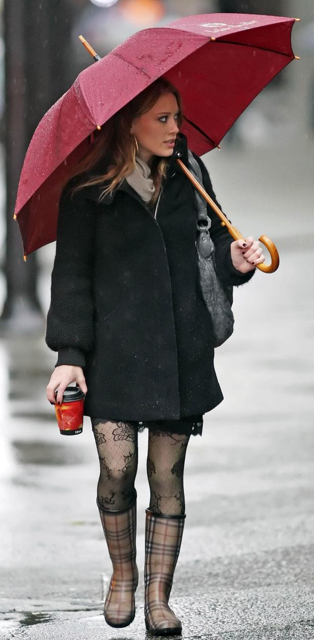 Eine Frau mit Regenschirm, Mantel und Gummistiefeln geht bei Regen die Straße entlang und hält einen Kaffeebecher.