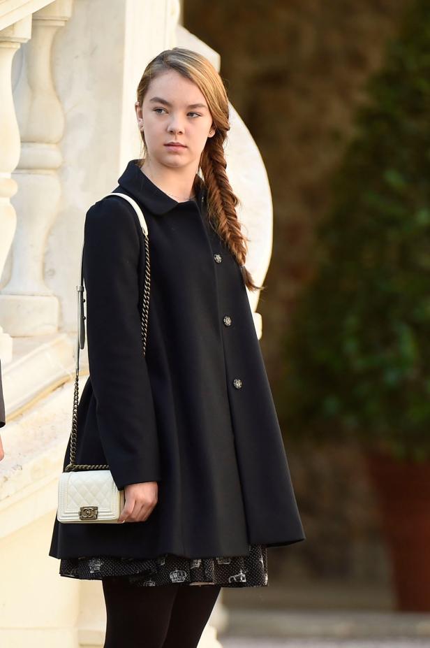 Ein junges Mädchen mit Zopf trägt einen schwarzen Mantel und eine kleine Handtasche.