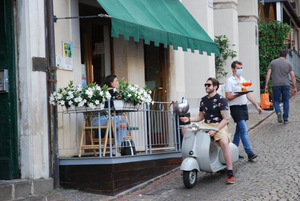 Ein Mann sitzt auf einem Roller vor einem Café, während ein Kellner Getränke serviert.