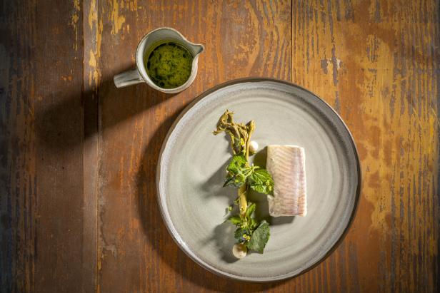 Ein Teller mit Fisch, Kräutern und einer Kanne grüner Sauce auf einem Holztisch.
