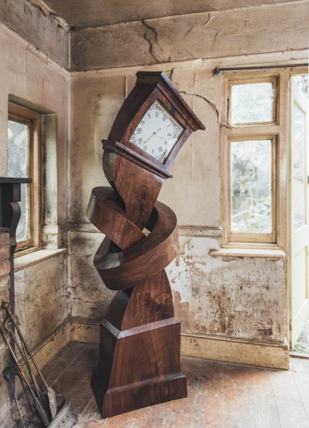 Eine skulpturale Standuhr aus dunklem Holz steht in einem Raum mit abblätternder Farbe.