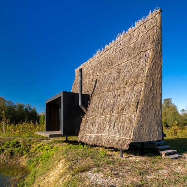 Ein kleines Haus mit Reetdach und einem schwarzen Anbau steht auf einer Wiese.