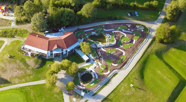 Luftaufnahme einer Minigolfanlage mit Teich und einem Gebäude mit rotem Dach.