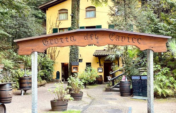 Der Eingang zum „Grotta del Cavicc“ mit einem hölzernen Schild und einem gelben Gebäude im Hintergrund.