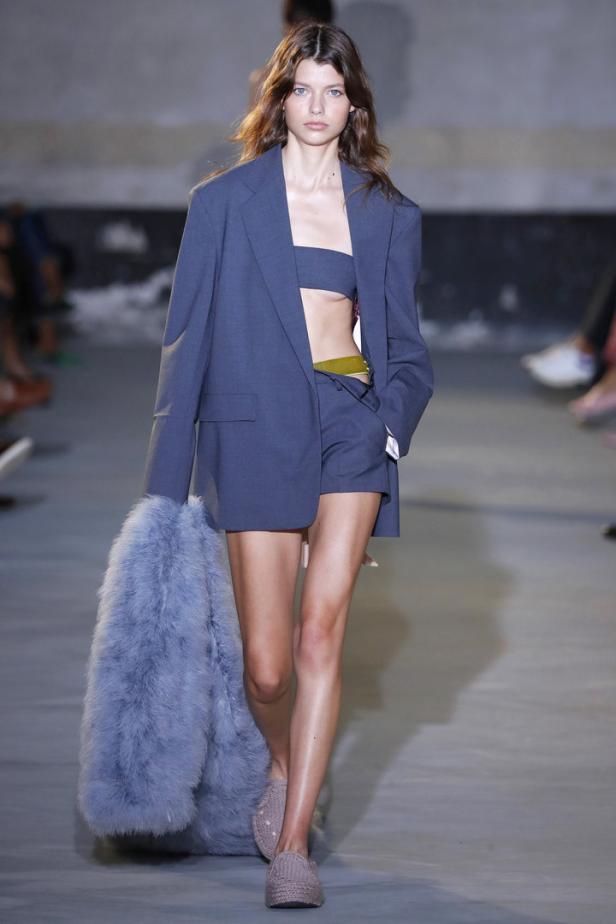 Ein Model auf dem Laufsteg, gekleidet in einen blauen Blazer, Shorts und ein kurzes Top, mit einem Pelzmantel über dem Arm.