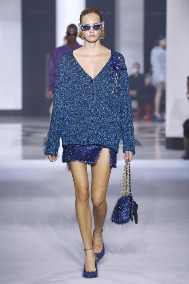 Ein Model in einem glitzernden, blaugrünen Cardigan und Minirock auf einem Laufsteg.
