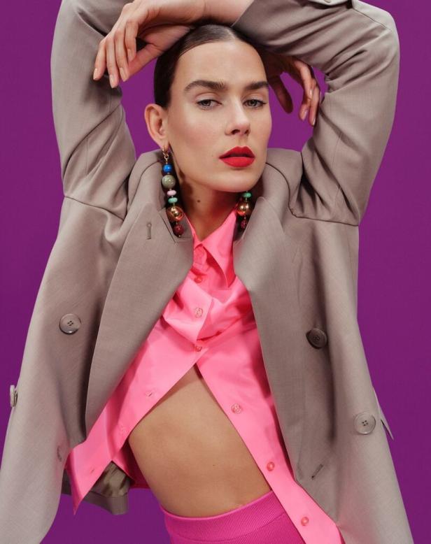 Eine Frau mit rotem Lippenstift trägt einen Blazer und eine pinkfarbene Bluse vor einem violetten Hintergrund.