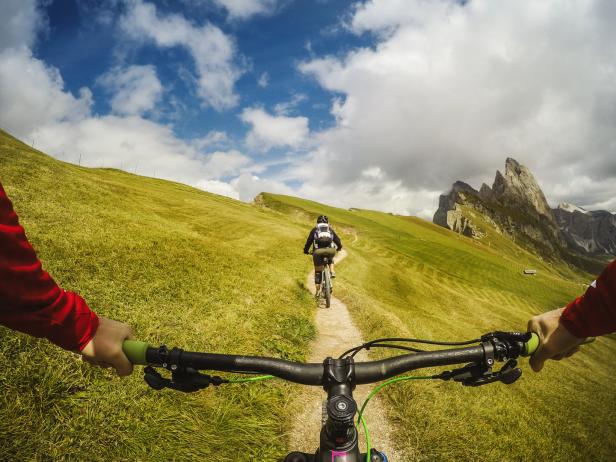 Mountainbiker fährt einen grasbewachsenen Pfad entlang, im Hintergrund die Geislerspitzen.