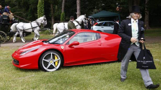 Ein roter Ferrari steht auf einer Wiese, im Hintergrund eine Kutsche mit weißen Pferden und ein Mann mit Zylinder.
