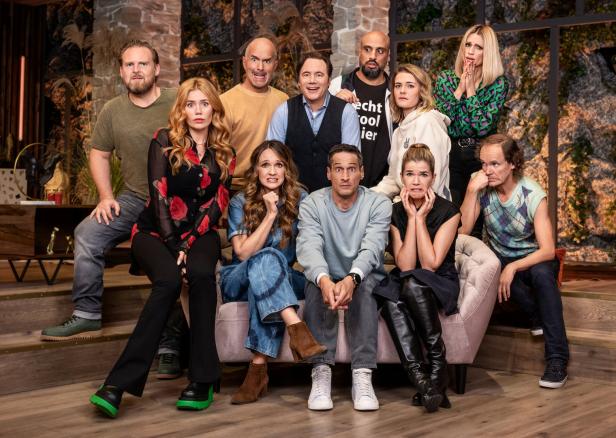Eine Gruppe von elf Personen sitzt auf einer Couch und blickt mit unterschiedlichen Gesichtsausdrücken in die Kamera.