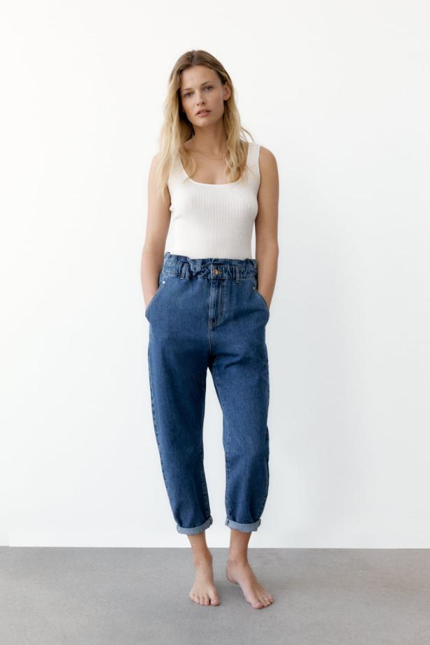 Eine Frau mit blonden Haaren trägt ein weißes Top und blaue Jeans.