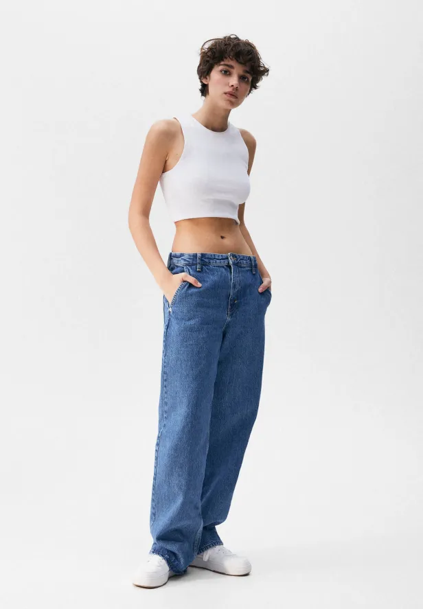Eine Frau mit kurzem Haar, gekleidet in Jeans und einem weißen Top, posiert vor einem weißen Hintergrund.