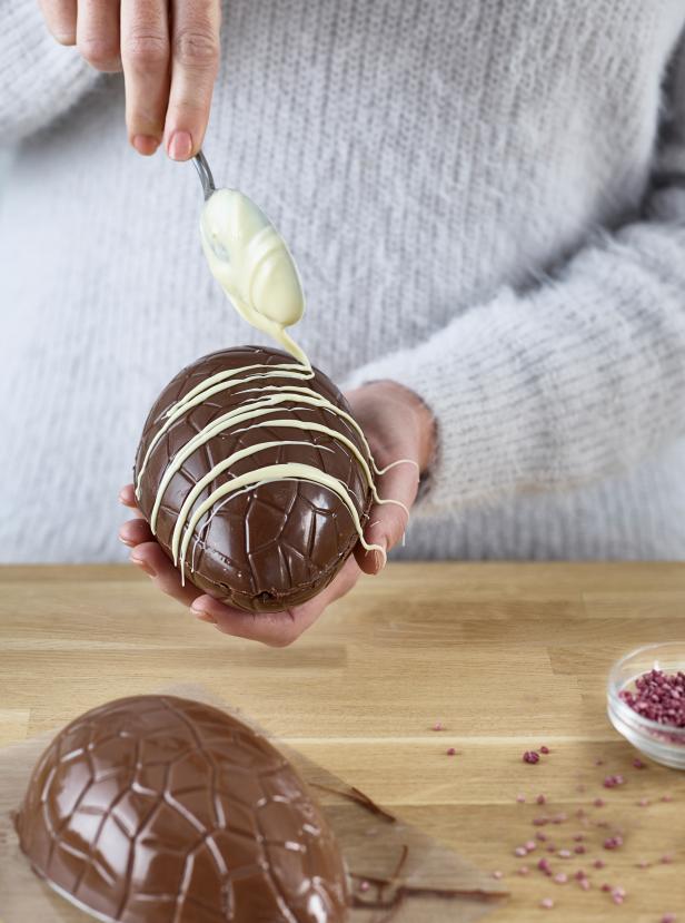 Eine Person dekoriert ein großes Schokoladenosterei mit weißer Schokolade.