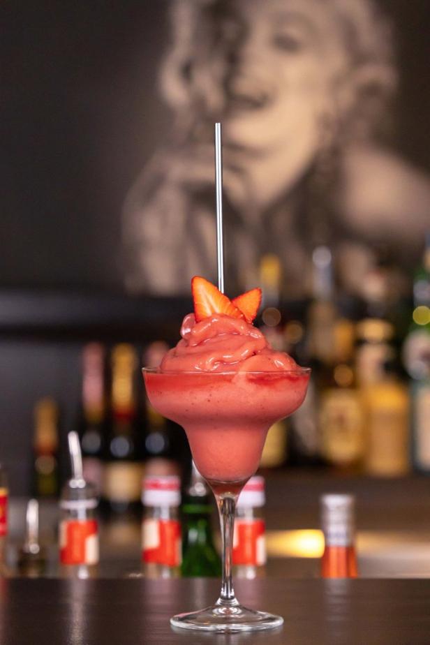 Ein Erdbeer-Daiquiri mit Strohhalm und Erdbeerstückchen vor einer Bar im Hintergrund.