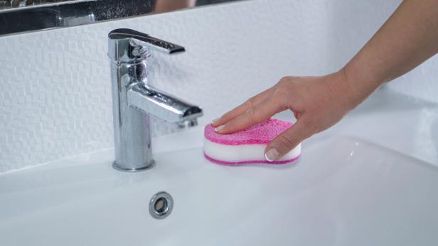 Eine Hand reinigt ein weißes Waschbecken mit einem rosa Schwamm.