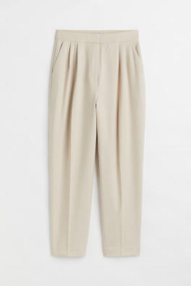 Eine hellbeige Stoffhose mit Bügelfalten hängt vor einem weißen Hintergrund.