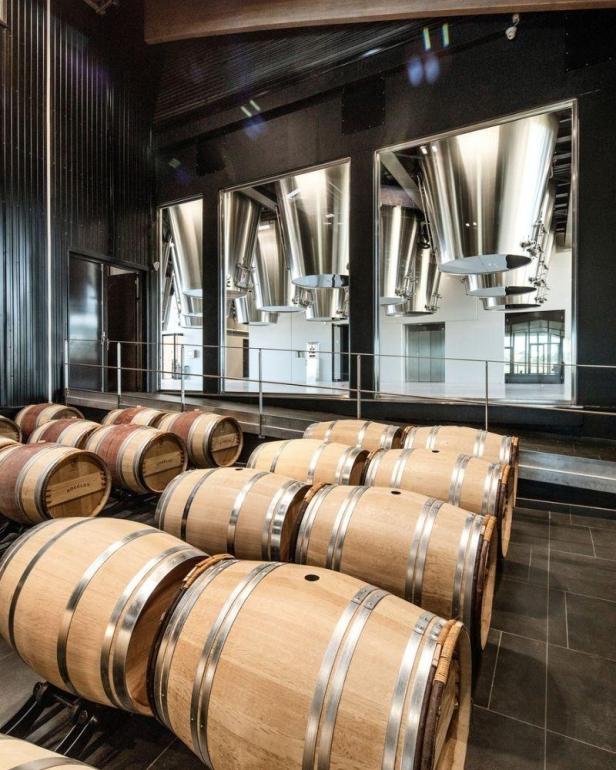 Mehrere Holzfässer stehen in einer modernen Lagerhalle mit Edelstahlbehältern.