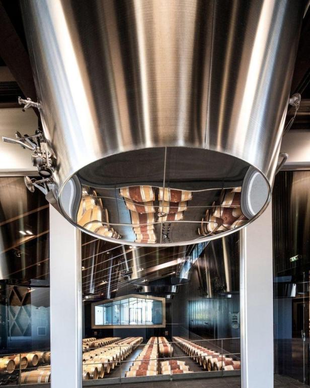 Ein Weinkeller mit Reihen von Fässern, die sich in einer großen, reflektierenden Metallstruktur spiegeln.