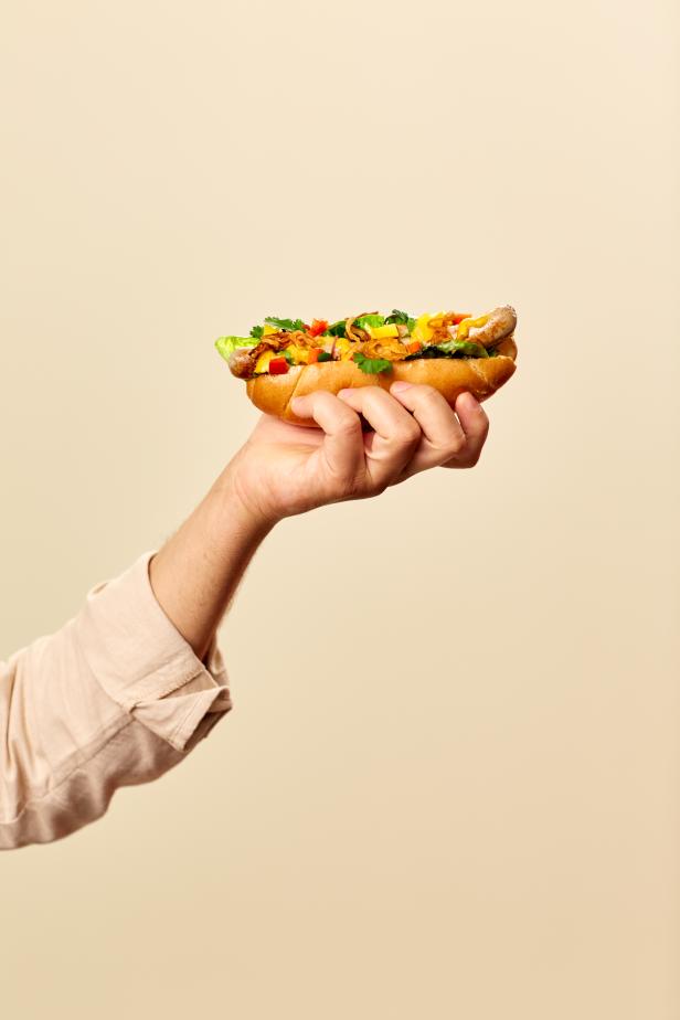 Eine Hand hält einen Hotdog mit verschiedenen Toppings vor einem beigen Hintergrund.