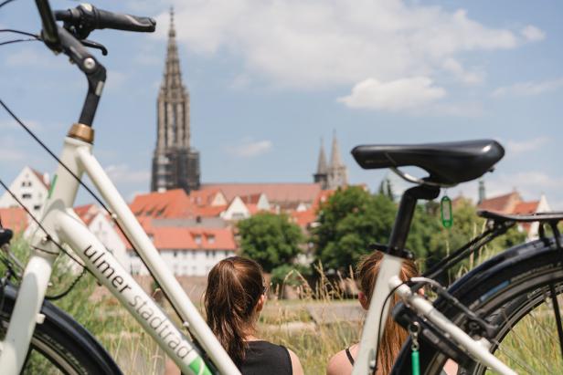 Zwei Frauen sitzen mit Fahrrädern im Vordergrund und blicken auf das Ulmer Münster.