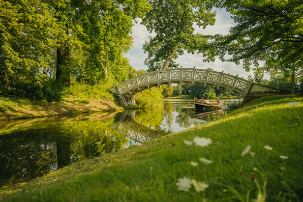 Park mit Bach und Brücke, Menschen rudern auf einem Boot unter der Brücke durch.
