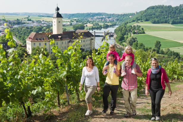 Eine Familie wandert durch einen Weinberg mit Blick auf ein Schloss und eine Flusslandschaft.