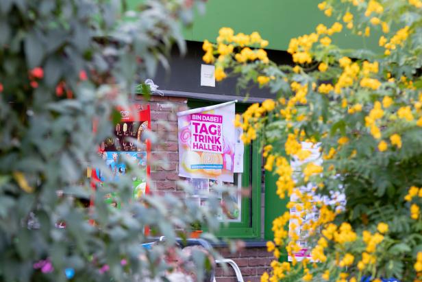 Ein Plakat für den „Tag der Trinkhallen“ hängt an einer Backsteinmauer, umgeben von gelben Blumen.