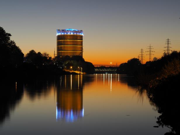 Ein beleuchteter Gasometer spiegelt sich im Wasser bei Sonnenuntergang.