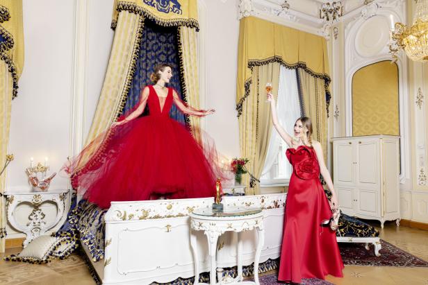 Zwei Frauen in roten Kleidern feiern in einem opulenten Schlafzimmer mit einem Glas Sekt.