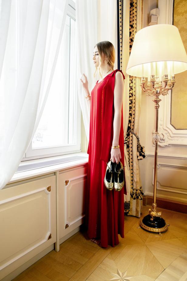 Eine Frau im roten Kleid blickt aus dem Fenster und hält goldene Schuhe in der Hand.