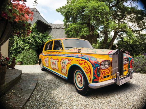 Ein gelber Rolls-Royce, verziert mit bunten Blumenmustern, steht vor einem Haus mit Strohdach.
