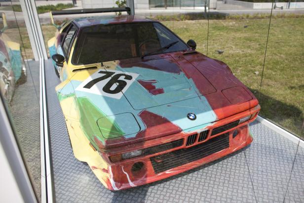 Ein bunt bemalter BMW M1 Art Car mit der Nummer 76 steht in einem gläsernen Ausstellungsraum.