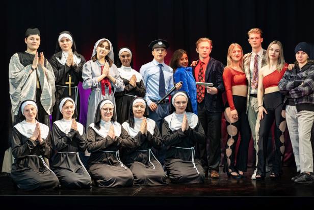 Eine Gruppe von als Nonnen, Polizist und Gangster verkleideten Personen steht auf einer Bühne.