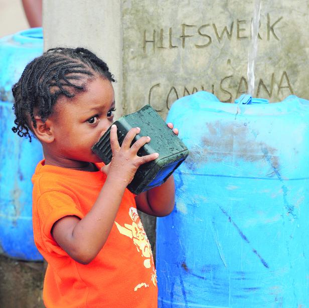 Ein kleines Mädchen trinkt Wasser aus einem Becher vor einem Hilfswerk-Brunnen.
