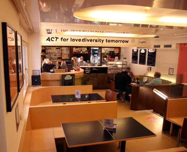 Innenansicht des „Magdas“-Cafés mit Gästen und dem Slogan „ACT for love diversity tomorrow“.