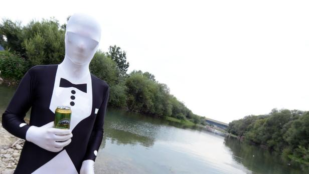 Eine Person in einem Ganzkörperanzug im Smoking-Design hält eine Dose Bier an einem Fluss.