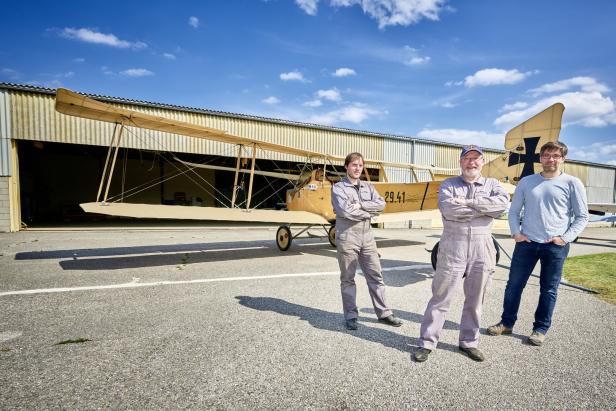 Drei Männer stehen vor einem historischen Flugzeug mit Hangar im Hintergrund.